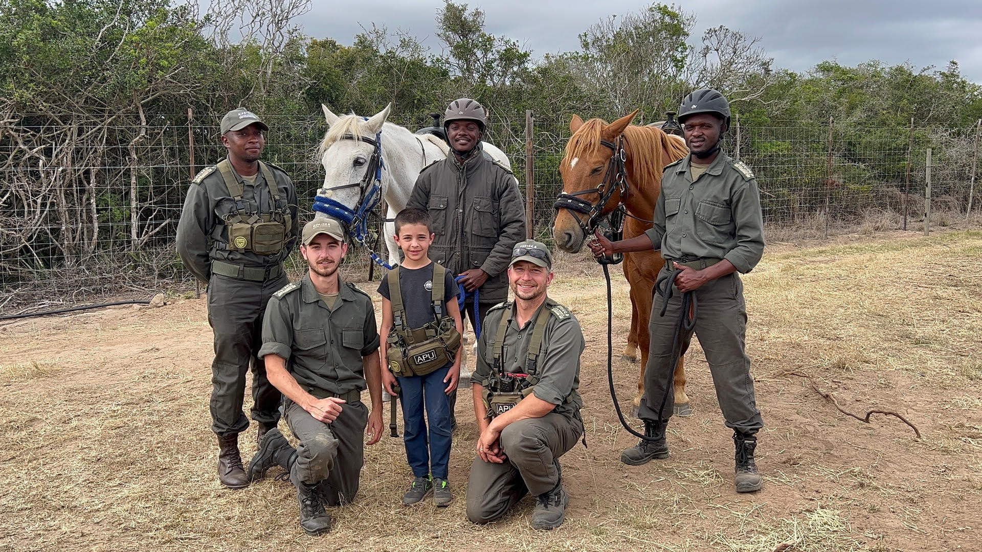 3 rangers standing with 2 horses and in front of them 2 rangers kneel on either side of a little boy