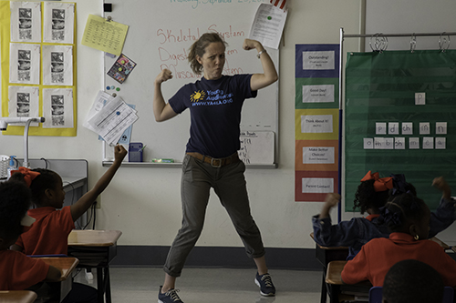 Teaching artist stands in front of the classroom holding up her muscles in an exaggerated stance.